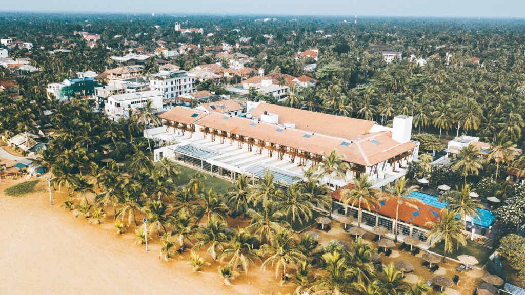 An outdoor elevated daytime view of Goldi Sands featuring a beachfront hotel building with an orange-tiled roof, swimming pool, palm trees, a sandy beach, and surrounding townscape in Sri Lanka