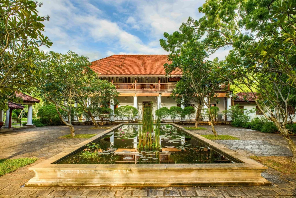 An outdoor daytime view of Ulagalla Walawwa featuring a historic manor house with an orange-tiled roof, a reflecting pond with water lilies in the courtyard, and lush tropical trees in Sri Lanka