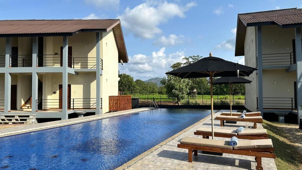 An outdoor daytime view of Hotel Sigiriya featuring a long swimming pool, wooden sun loungers with black umbrellas, multi-story hotel buildings with balconies, and surrounding greenery under a bright sky in Sri Lanka