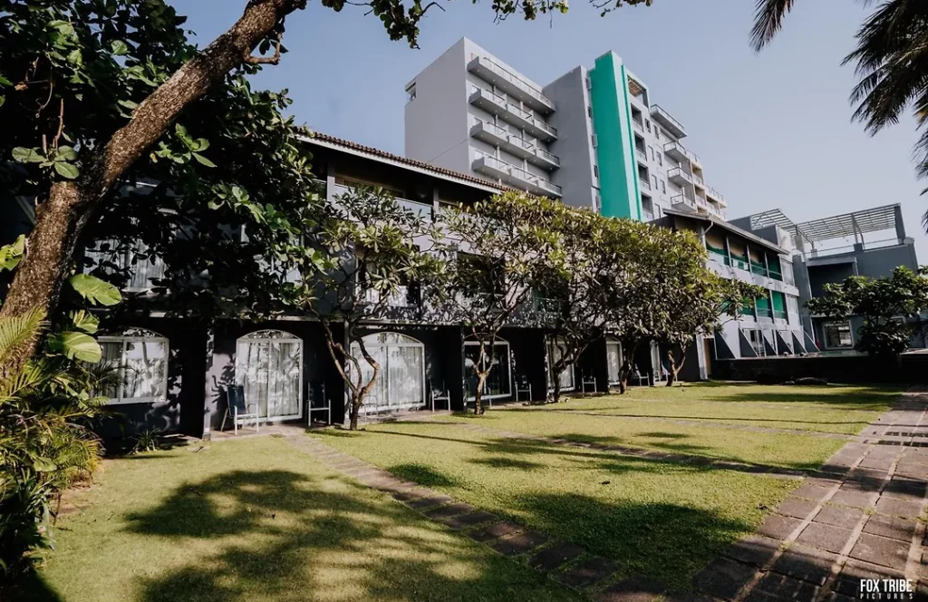 An outdoor daytime view of Lali's resort featuring a modern low-rise grey hotel building with a manicured green lawn, stone pathways, mature trees, and a taller white and teal tower block in the background in Sri Lanka