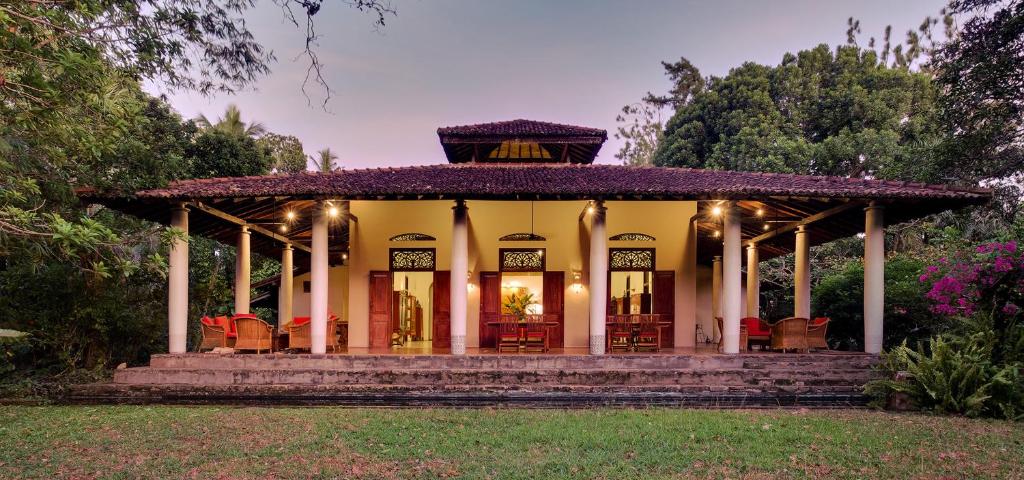 An outdoor twilight view of Apa Villa in Thalpe featuring the warmly lit colonial-style pillared veranda and red-tiled roof surrounded by tropical gardens in Sri Lanka