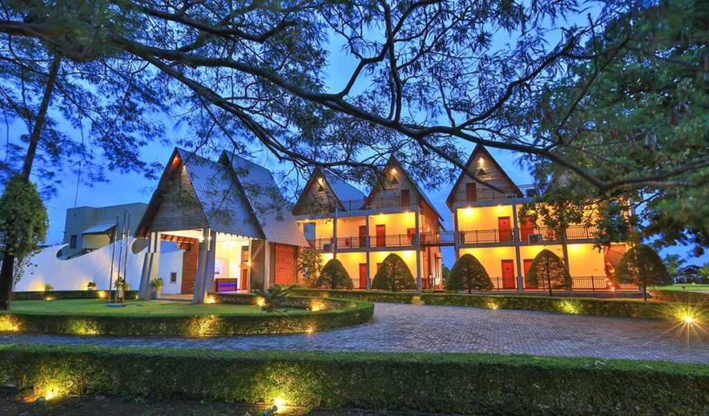 An outdoor evening twilight view of Lava Waves featuring illuminated resort chalets with pitched roofs, warm ground lighting, and manicured landscaping framed by tree branches in Sri Lanka