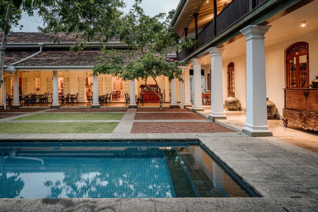 An outdoor daytime view of Maniumpathy Hotel in Colombo, featuring a sparkling lap pool, classic white pillars, and the restored heritage mansion facade set within Cinnamon Gardens, Sri Lanka