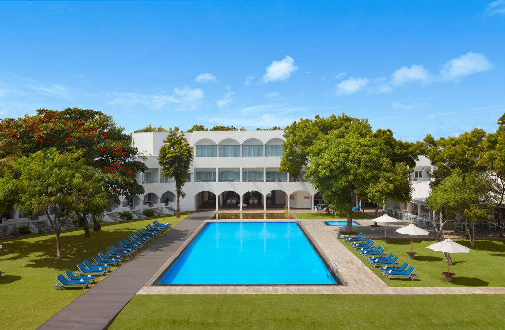 An outdoor daytime view of Trinco Blu featuring a large swimming pool, neat rows of blue sun loungers on a green lawn, and white resort buildings under a bright blue sky in Sri Lanka
