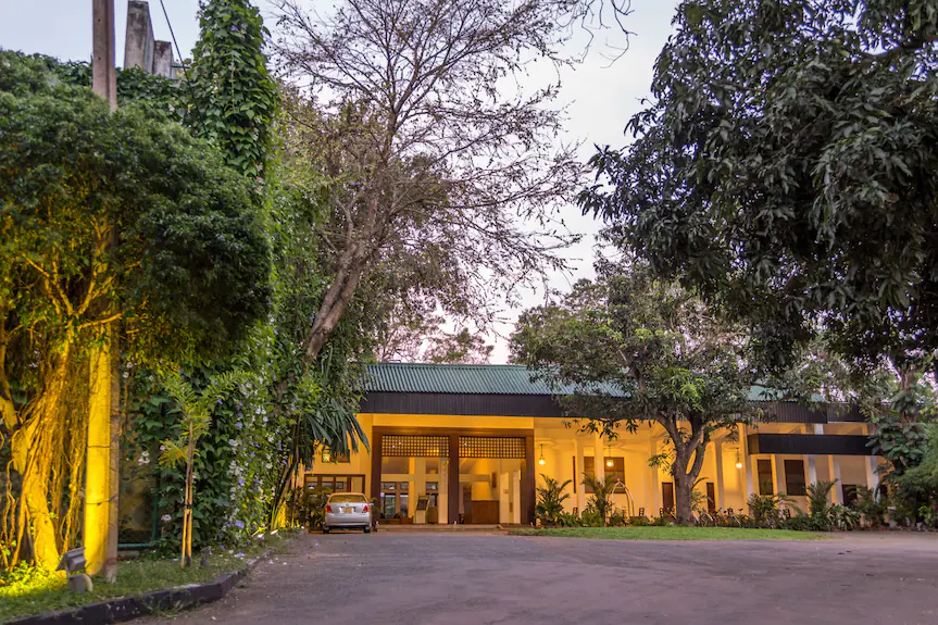 An outdoor evening view of Lake Side by Nuwara Wewa featuring a illuminated hotel building entrance, a paved driveway, a parked car, and lush surrounding trees under a twilight sky in Sri Lanka