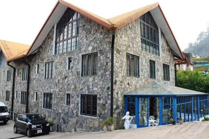 An outdoor daytime view of a multi-story stone exterior building with a pitched orange-tiled roof, multiple windows, a blue-framed sunroom extension, and parked cars in Sri Lanka
