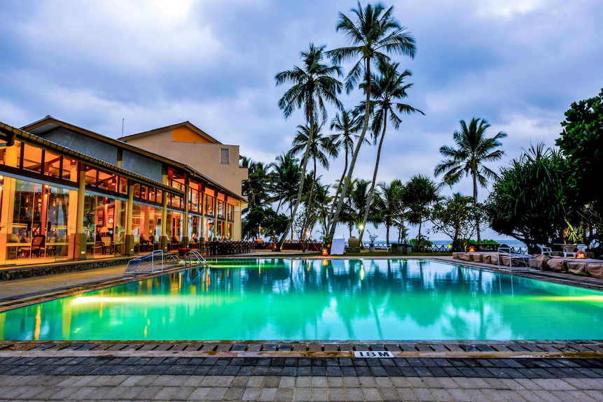 An outdoor twilight view of Insight Hotel featuring an illuminated swimming pool, resort buildings, and tall palm trees framing the oceanfront in Ahangama, Sri Lanka