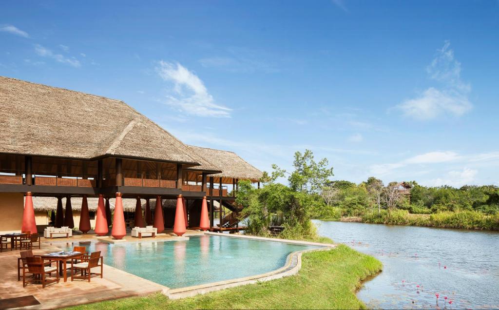 An outdoor daytime view of Vil Uyana featuring luxury thatched-roof wooden chalets, an outdoor swimming pool, a dining terrace, and a tranquil water body under a bright blue sky in Sri Lanka