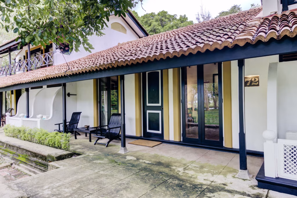An outdoor daytime view of Cinnamon Lodge Habarana featuring a traditional guest room exterior with a terracotta-tiled pitched roof, a covered veranda with black outdoor seating, and a stone walkway in Sri Lanka