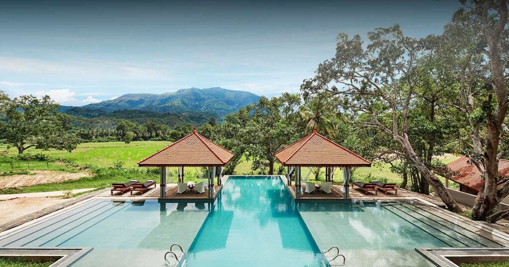 An outdoor high-angle daytime view of Jetwing Kaduruketha featuring an outdoor swimming pool with pavilion gazebos, overlooking working paddy fields and majestic mountains in Wellawaya, Sri Lanka