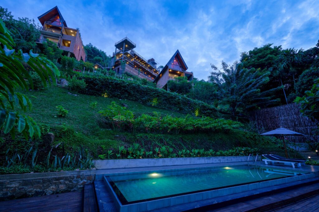 An outdoor twilight view of Majentra, an upscale boutique escape offering scenic hillside views, modern amenities, an illuminated swimming pool, and lush terraced greenery in Ella, Sri Lanka