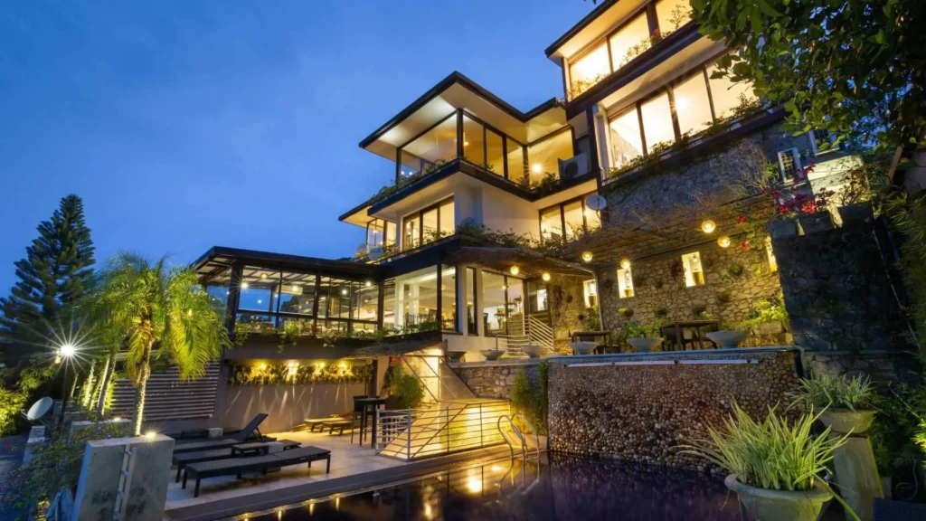 An outdoor twilight view of Theva Residency boutique hotel perched on the Hanthana slopes, featuring a lighted infinity pool, stone walls, and multi-level glass architecture in Kandy, Sri Lanka