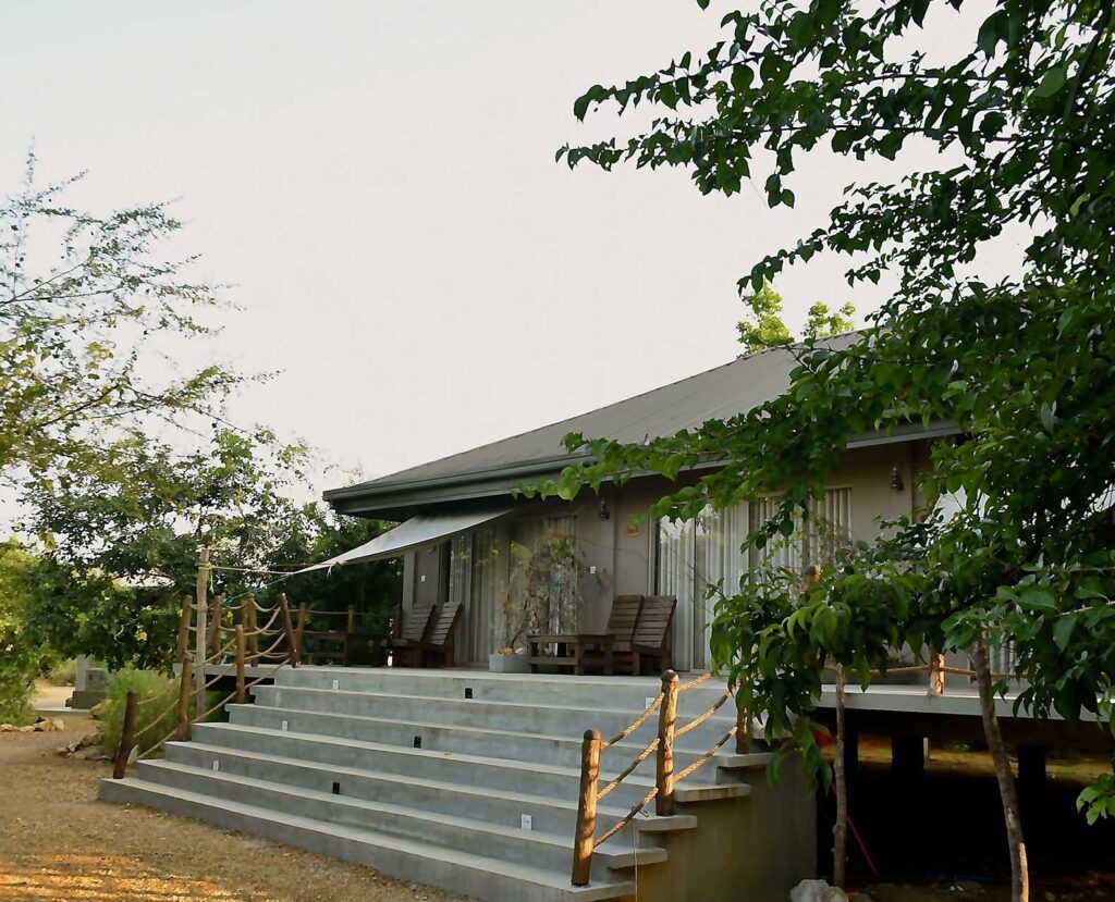 An outdoor daytime view of Kale Resort, a cozy, nature-focused lodge featuring a raised chalet with concrete steps, rustic wooden railings, and surrounding wilderness near Yala, Sri Lanka