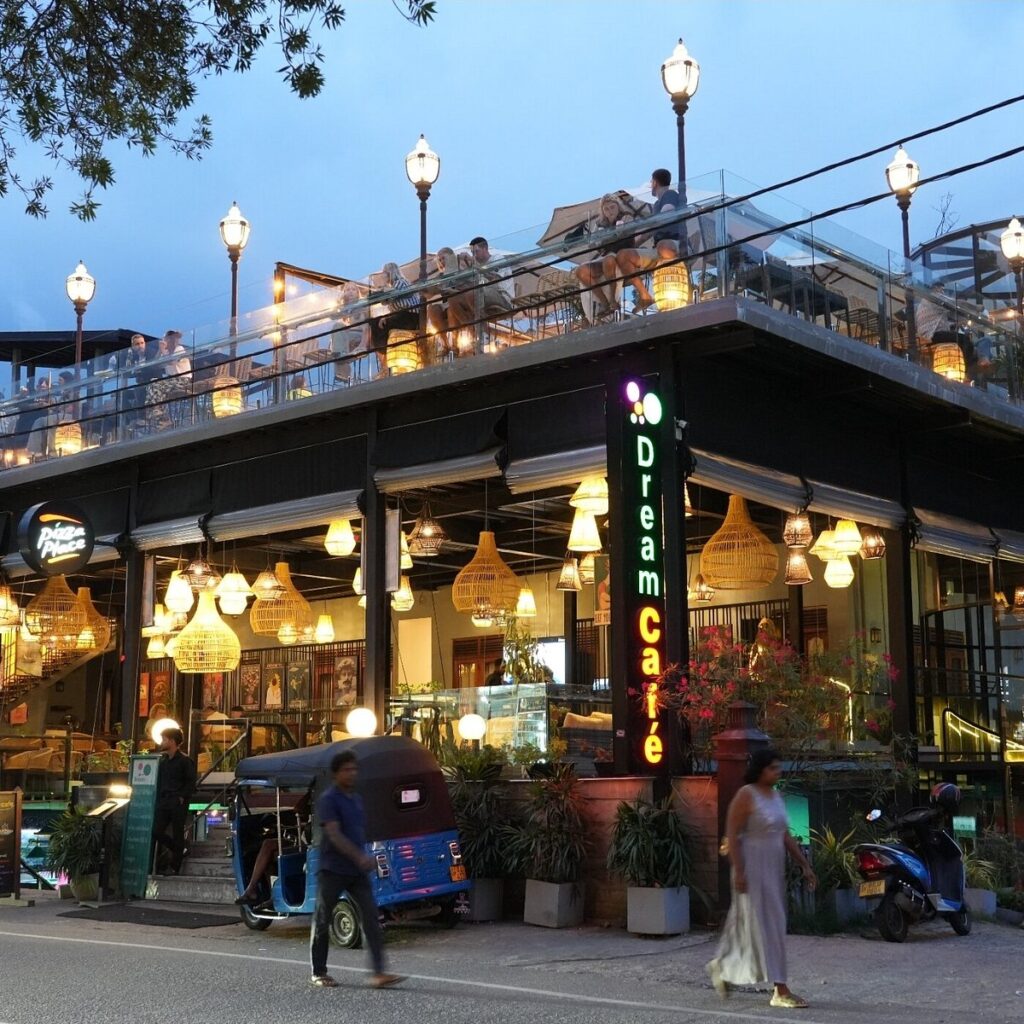 An outdoor evening street-level view of Dream Cafe featuring a multi-level building with glowing warm lights, a rooftop terrace dining area, and pedestrians on the street in Ella, Sri Lanka