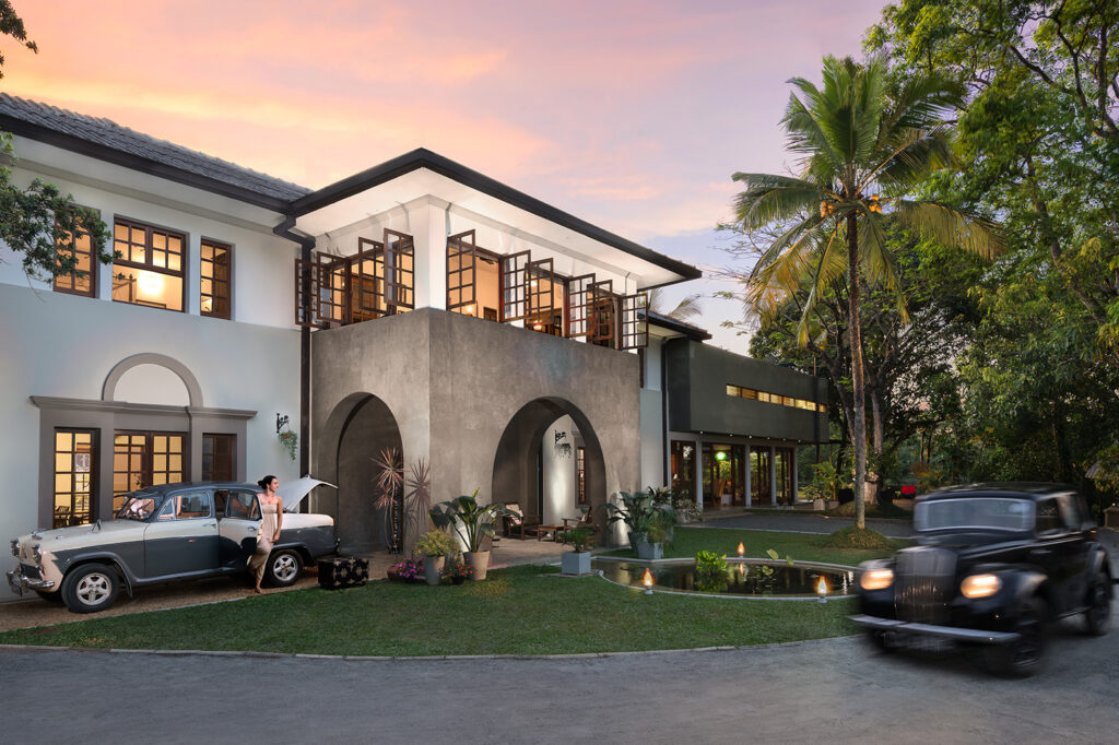An outdoor twilight view of Elephant Stables featuring classic colonial architecture, arched entryways, vintage cars parked on the driveway, and palm trees under a warm sunset sky in Sri Lanka