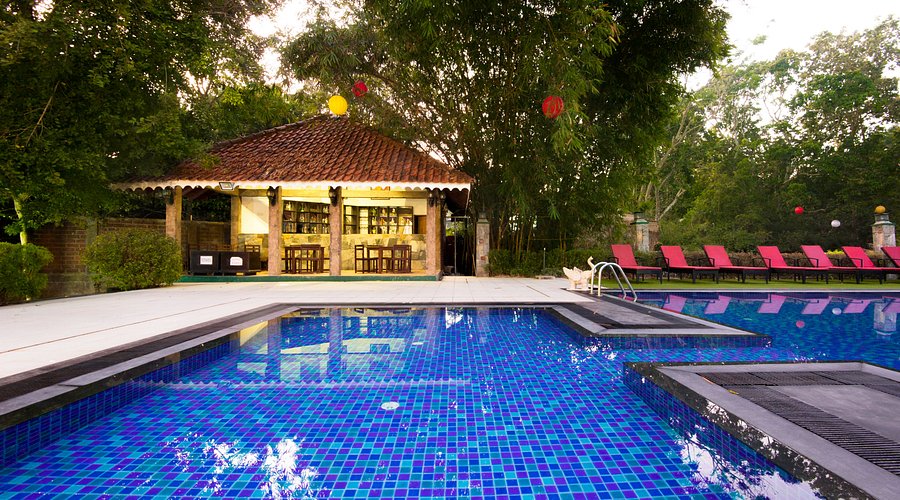 An outdoor daytime view of Heritage Hotel Anuradhapura featuring a vibrant blue-tiled swimming pool, a line of red sun loungers, a pavilion with a traditional tiled roof, and lush trees in Sri Lanka