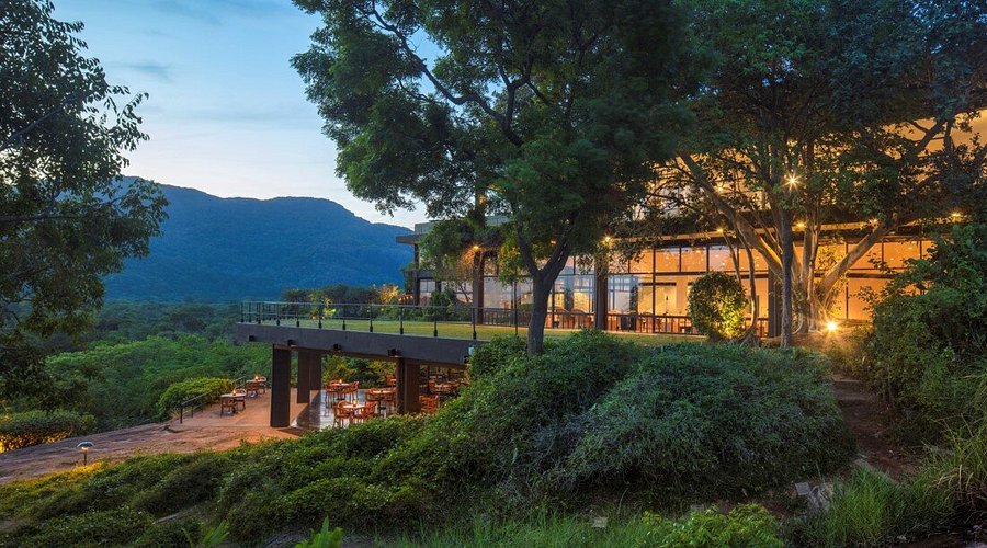 An outdoor evening twilight view of Heritance Kandalama featuring a modern resort building embedded into lush green hills, warm interior lights, an elevated terrace, and a scenic mountain backdrop in Sri Lanka