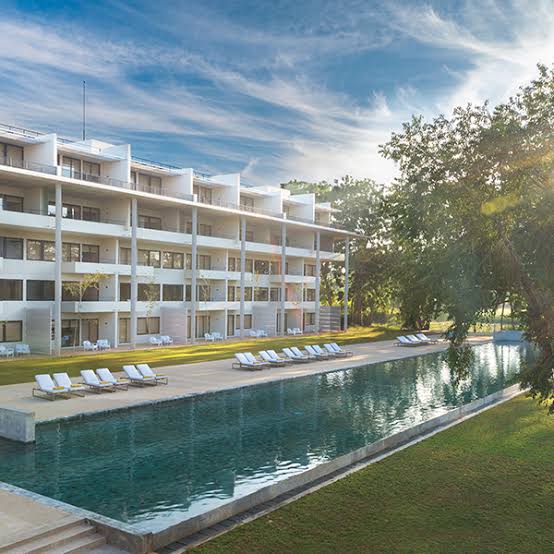 An outdoor daytime view of Jet Wing Lake featuring a modern multi-story resort hotel building, a long swimming pool with sun loungers, green lawns, and surrounding trees under a bright blue sky in Sri Lanka