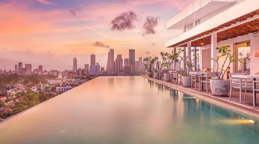 An outdoor rooftop sunset view of Jetwing Colombo 7 featuring an infinity pool and bar terrace overlooking the glowing city skyline of Colombo, Sri Lanka