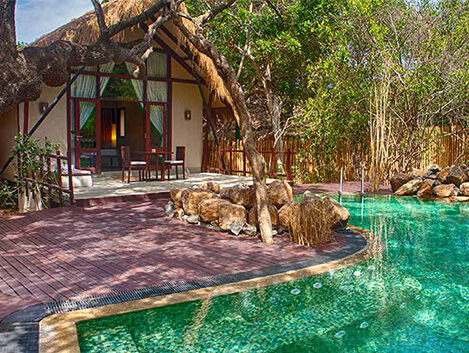 An outdoor daytime view of Jungle Beach by Uga featuring a rustic wooden resort villa exterior, a curved swimming pool, and dense surrounding jungle trees in Sri Lanka