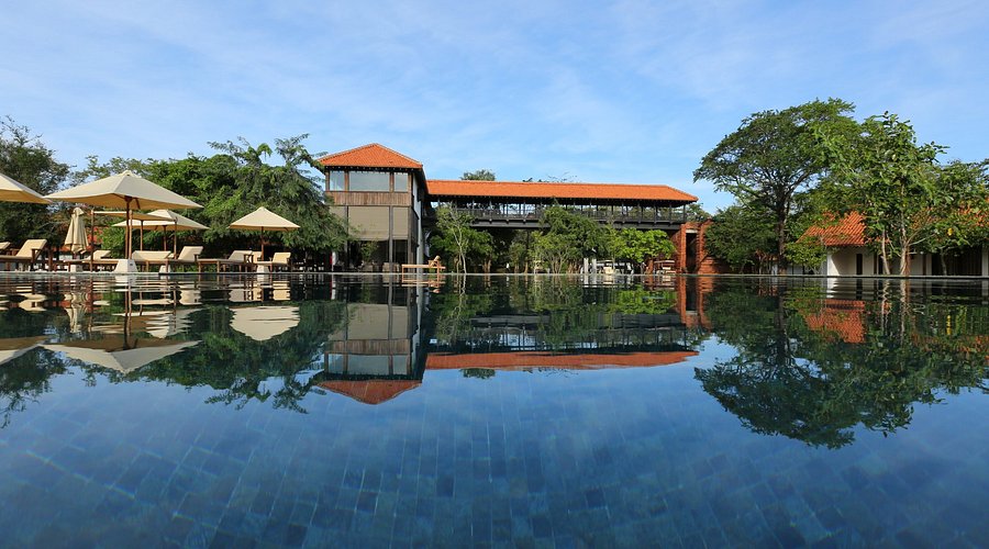 An outdoor daytime view of Sigiriya Jungle featuring a large infinity swimming pool reflecting the resort buildings, sun umbrellas, and surrounding green trees under a clear blue sky in Sri Lanka