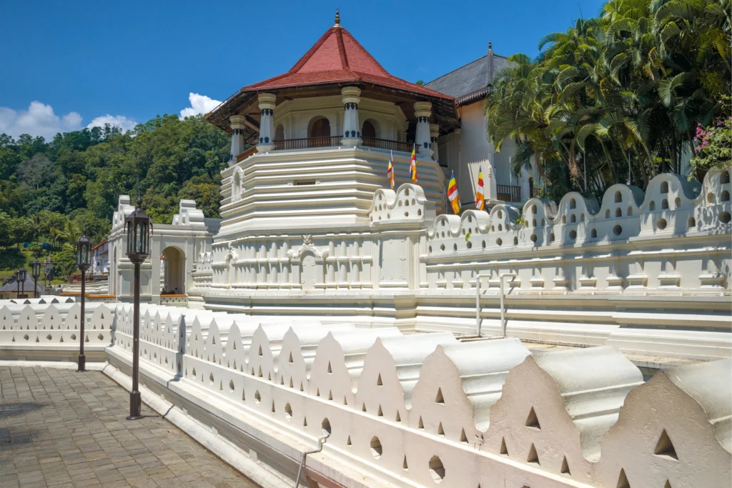 An outdoor daytime view in Sri Lanka featuring the white ornate walls and multi-tiered red-tiled roof of the Temple of the Sacred Tooth Relic in Kandy against a bright blue sky