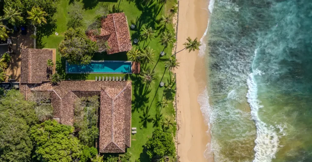 An outdoor aerial daytime view of The Frangipani Tree in Thalpe, featuring red-roofed buildings, a swimming pool, green lawns, and a sandy beach meeting the ocean waves in Sri Lanka