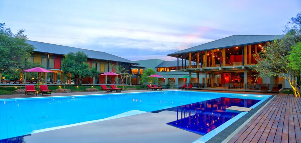 An outdoor eye-level dusk view of an eco-conscious resort in Dambulla featuring a large central swimming pool surrounded by a wooden deck, loungers, and lush garden greenery under an evening sky