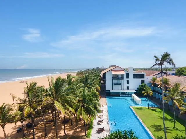 An outdoor elevated daytime view of Jetwing Blue featuring a beachfront resort with a long swimming pool, palm trees, a sandy beach, and the ocean under a bright blue sky in Sri Lanka